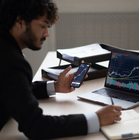 Smart focused Indian broker investor, stock trader using smartphone and laptop to analyze and invert in the financial market of cryptocurrencies, buy or sell crypto coins, planning strategy