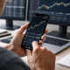 A trader analyzes a financial price chart on a smartphone while multiple market charts display on monitors in the background.